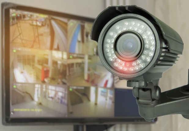 Picture of a monitor showing four video feeds from security cameras beside a security camera that has a red light to indicate it is recording.
