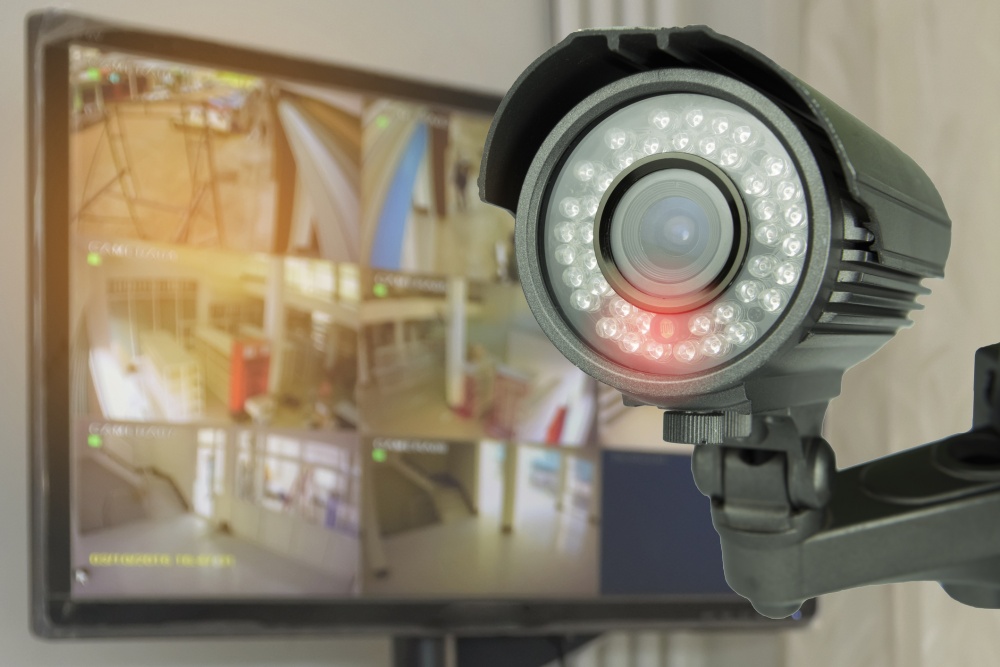 Picture of a monitor showing four video feeds from security cameras beside a security camera that has a red light to indicate it is recording.